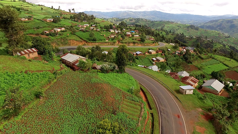 A winding road cuts through a lush, hilly landscape dotted with houses and patches of agricultural fields under a cloudy sky.