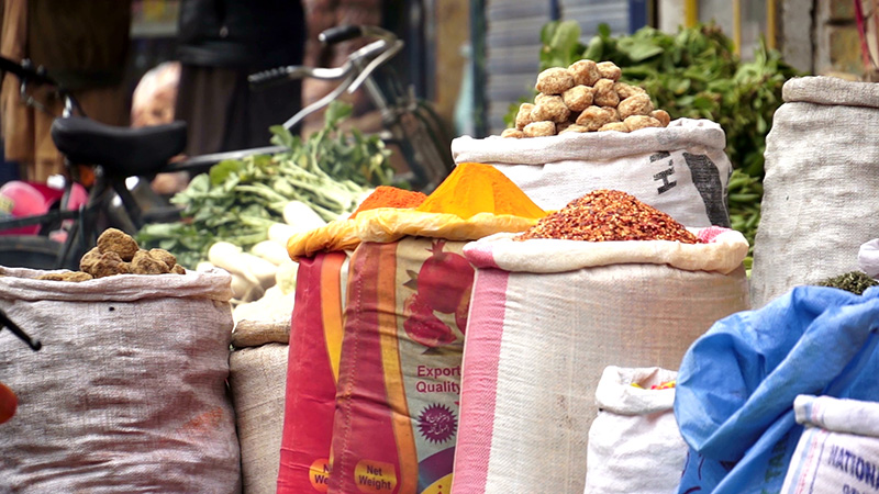 Sacks of various grains and legumes, vividly colored spices, and fresh green vegetables displayed for sale in a market setting.