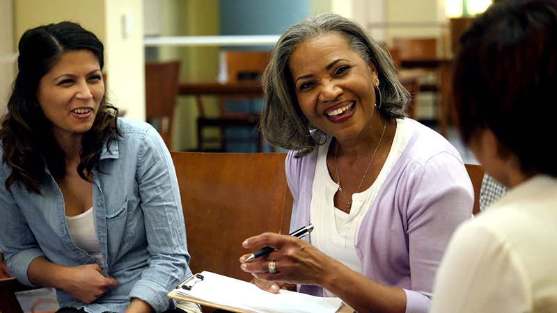 Three people in a conversation, one of whom is holding a pen and paper, with expressions of engagement and happiness. The setting appears to be an indoor environment conducive to discussion or learning.