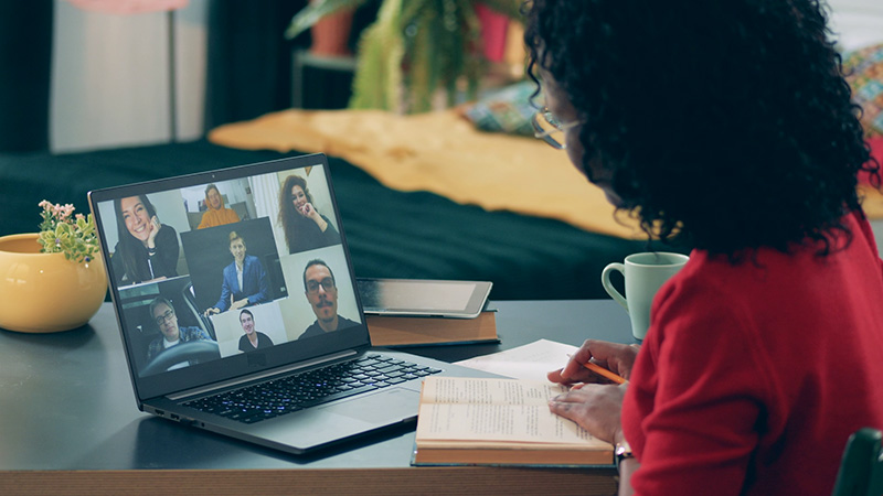 A person in a red shirt is participating in a virtual meeting with seven other individuals on a laptop screen, sitting at a desk with a notebook, pen, and a green mug. There is a yellow pot with flowers beside the laptop.