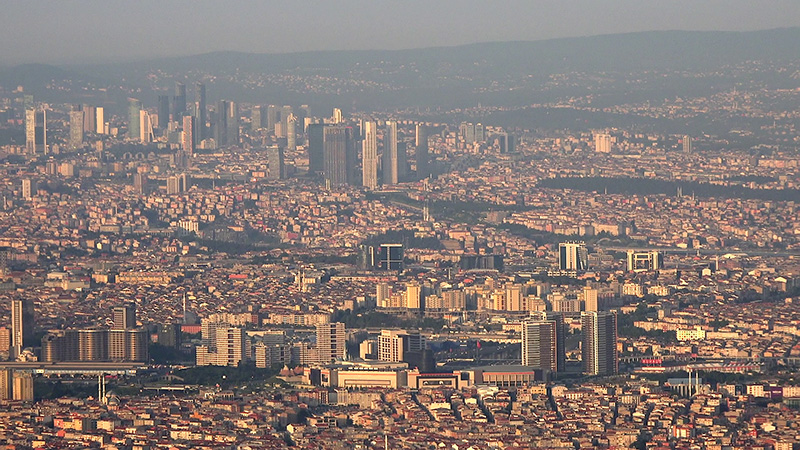 Aerial view of a dense cityscape, with numerous buildings packed closely together, high-rises in the distance.