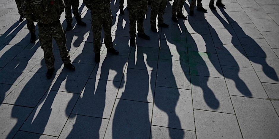A group of people in army uniforms stand on concrete, there are long shadows on the ground.
