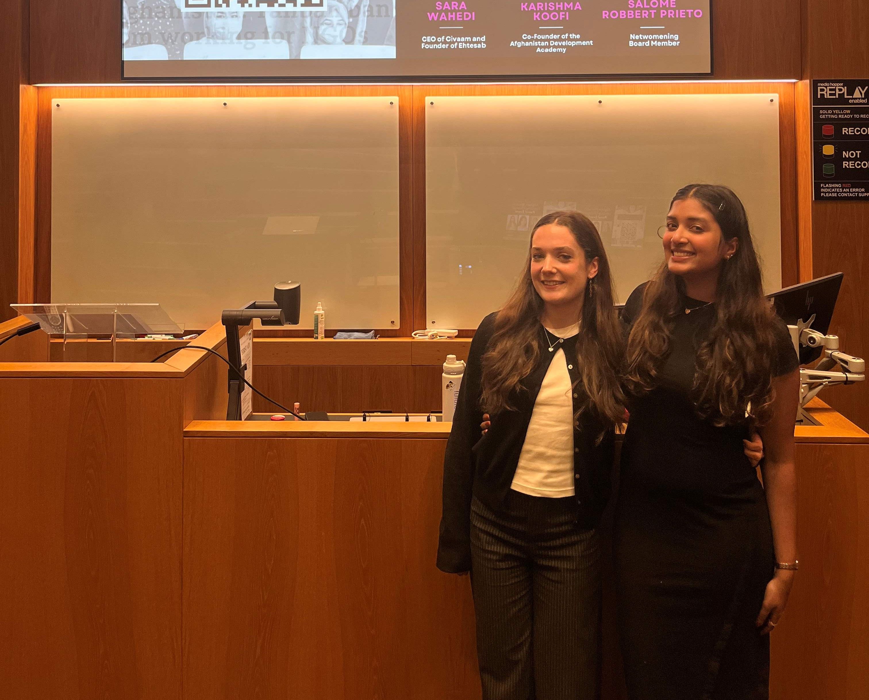 Photo of Thulsa Miqdhaadh Moosa and a friend standing at the front of a lecture hall. Behind them is a screen that reads 'Afghan Women Leading Panel Change: Panel Event'