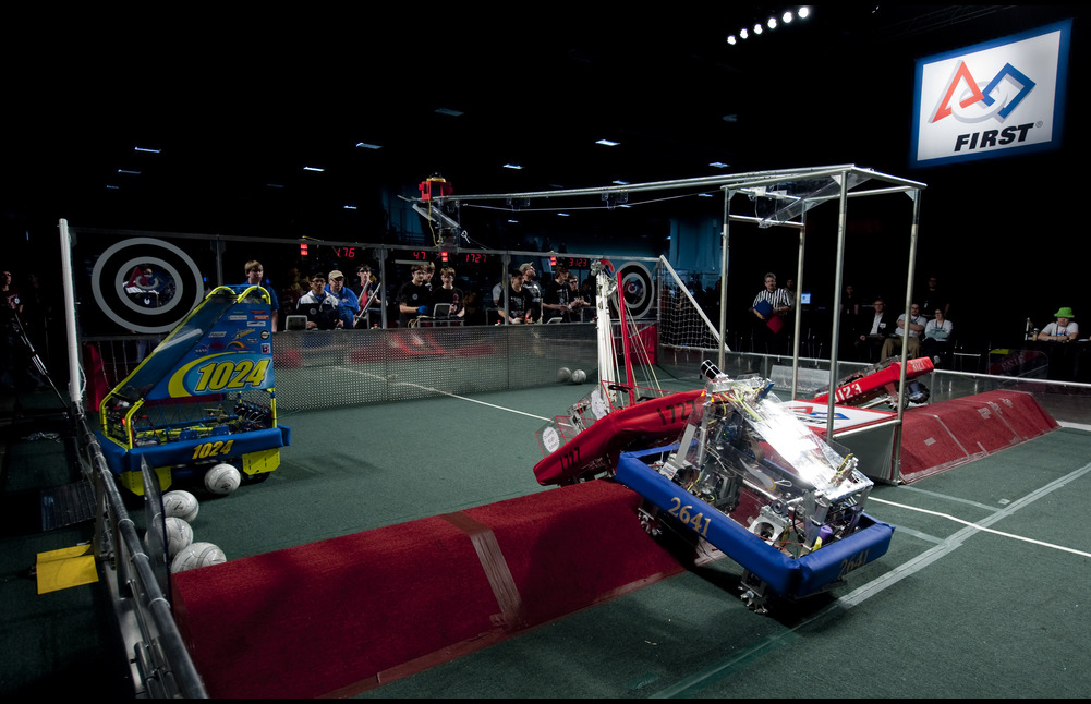 Students operate blue, red and white coloured robotic machinery in a competitive setting.
