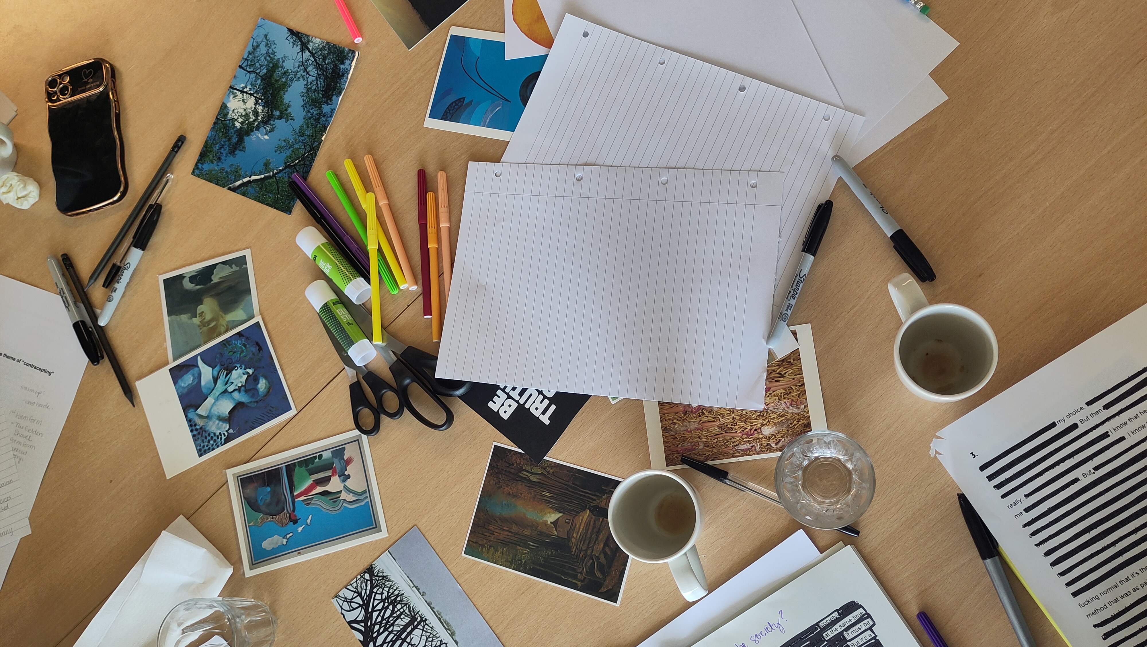 Table with postcards, lined paper, pens, scissors, tea and water, and a halfwritten poem