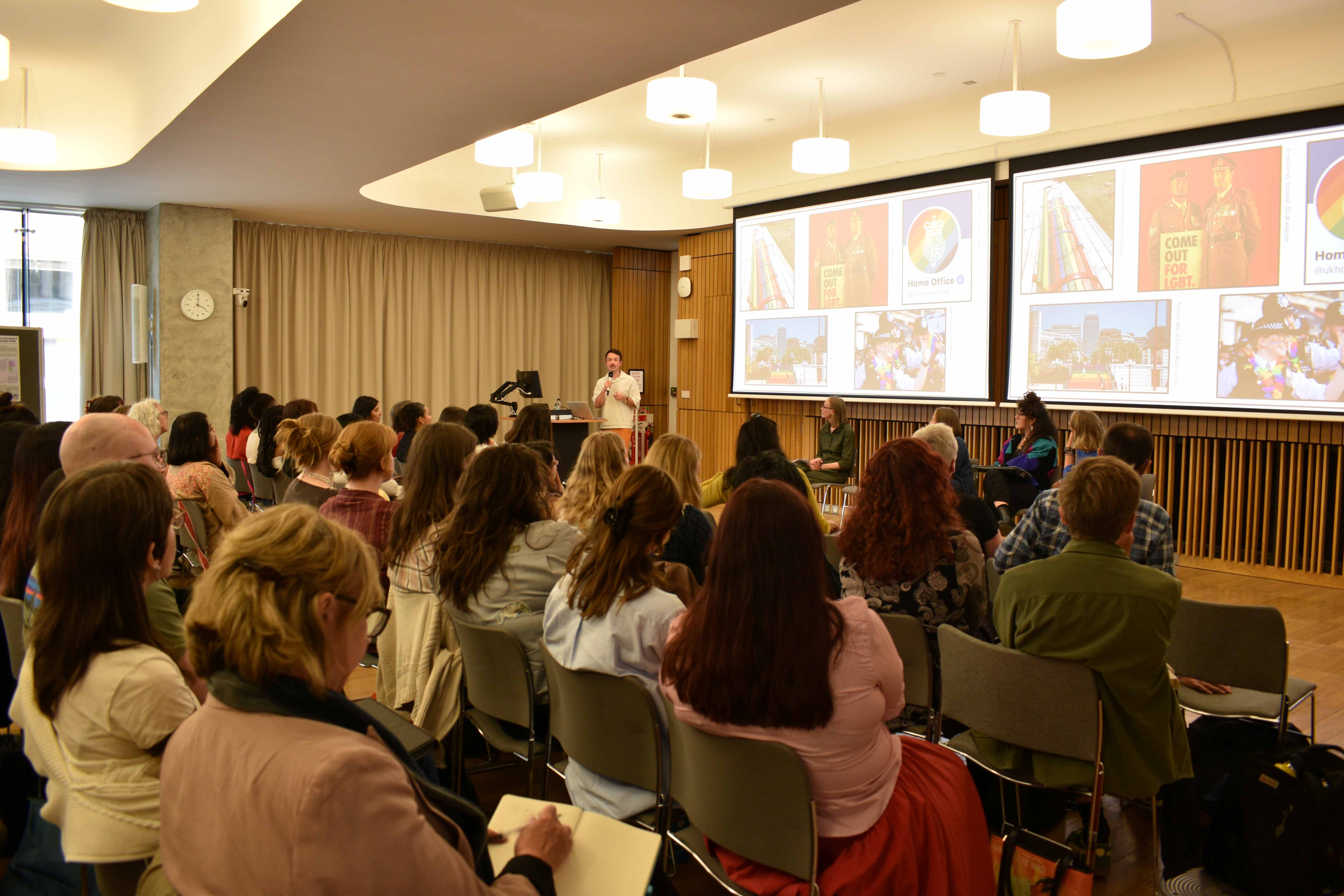 Photo of Kevin Guyan giving his presentation as part of the panel discussion. He is holding a micrphone. To his left, there is a slideshow and the rest of the panelists are sitting.