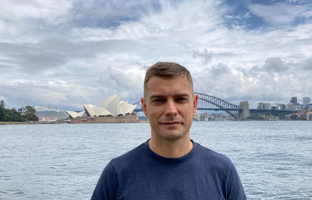Man smiling in front of a water body with a bridge in the background