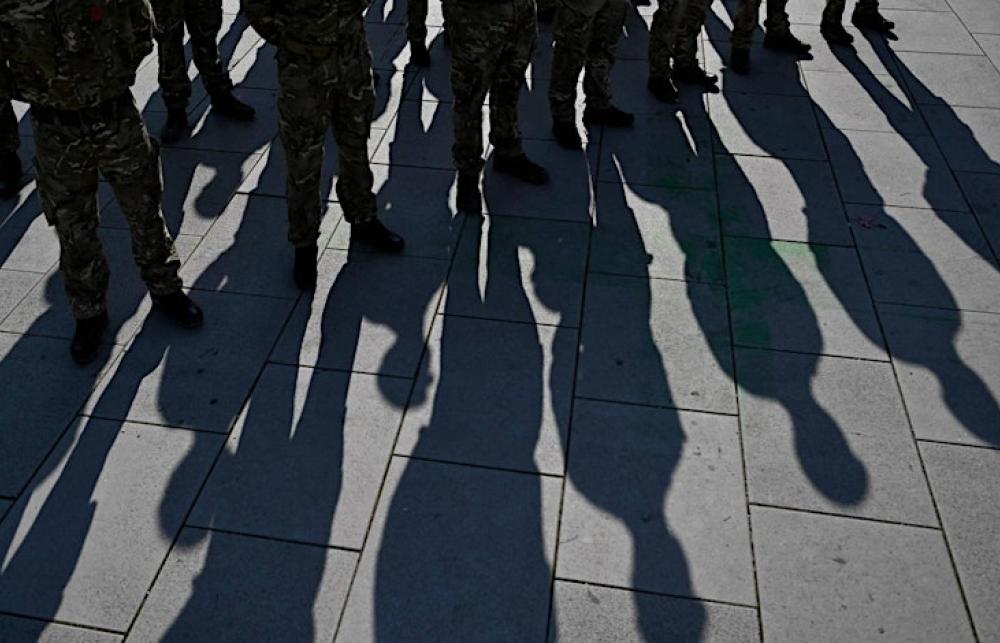 A group of people in army uniforms stand on concrete, there are long shadows on the ground.
