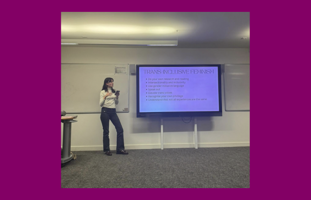 Photo of Ione Gildroy standing next to a screen that reads 'Trans Inclusive Feminism' and a list of bullet points on how to act in accordance with trans inclusive feminism. 