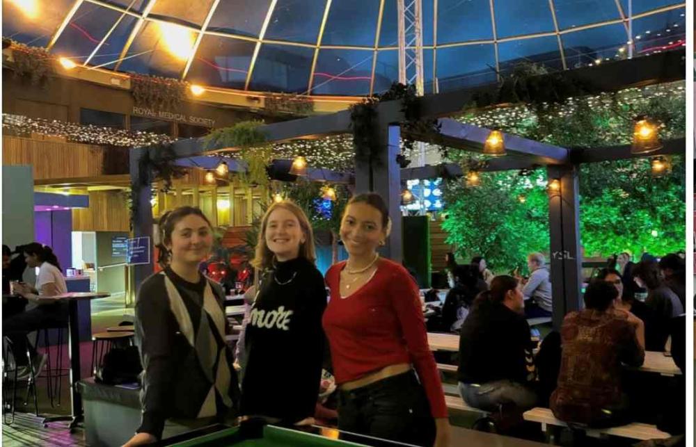 A photo of Mia Hiraani-Nelson with two frinds by a pool table in Potterow, the University of Edinburgh Student Union. Behind them is a dome and a lot of people socializing on benches. 