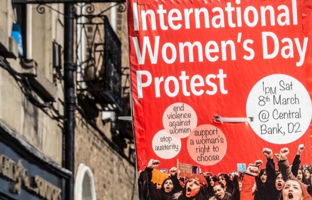 Photo of a poster on a lamp post in a street that reads 'International Women's Day Protest'. There is an image of people protesting and chanting under the text on the poster.