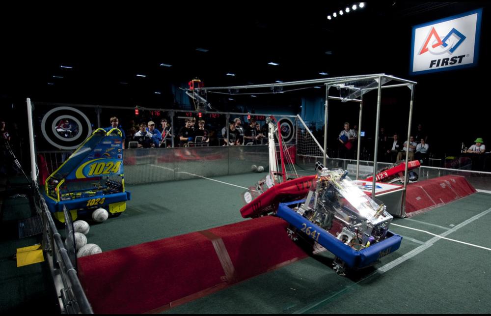 Students operate blue, red and white coloured robotic machinery in a competitive setting.