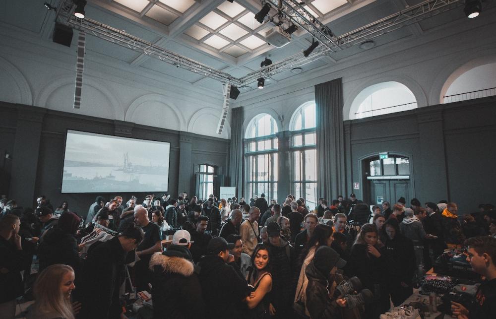 A spacious room filled with people. A presentation plays on a projector screen.